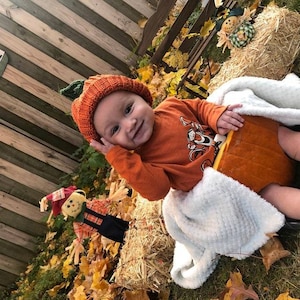 Puede incluir: Un beb&eacute; disfrazado de calabaza naranja, con un gorro de punto y un tallo verde. El beb&eacute; lleva una camiseta naranja de manga larga con un personaje de dibujos animados. El beb&eacute; est&aacute; dentro de una calabaza tallada y envuelto en una manta blanca.