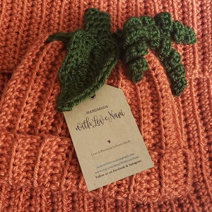 Puede incluir: Primer plano de una calabaza de ganchillo naranja hecha a mano con hojas verdes. La calabaza tiene una textura acanalada y una etiqueta que dice "HANDMADE With Love Nani". La etiqueta tambi&eacute;n incluye informaci&oacute;n de contacto.