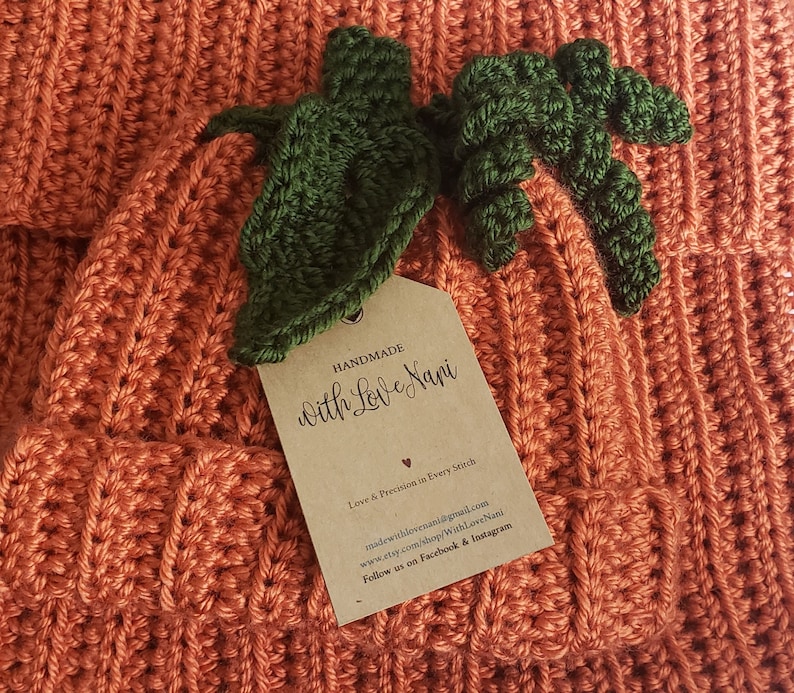 Puede incluir: Primer plano de una calabaza de ganchillo naranja hecha a mano con hojas verdes. La calabaza tiene una textura acanalada y una etiqueta que dice "HANDMADE With Love Nani". La etiqueta tambi&eacute;n incluye informaci&oacute;n de contacto.