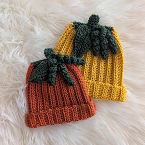Puede incluir: Dos gorros de ganchillo hechos a mano con forma de calabaza. Uno es naranja, el otro amarillo. Ambos tienen detalles de hojas y tallos verdes. Los gorros son acanalados y se asientan sobre una superficie blanca y esponjosa.