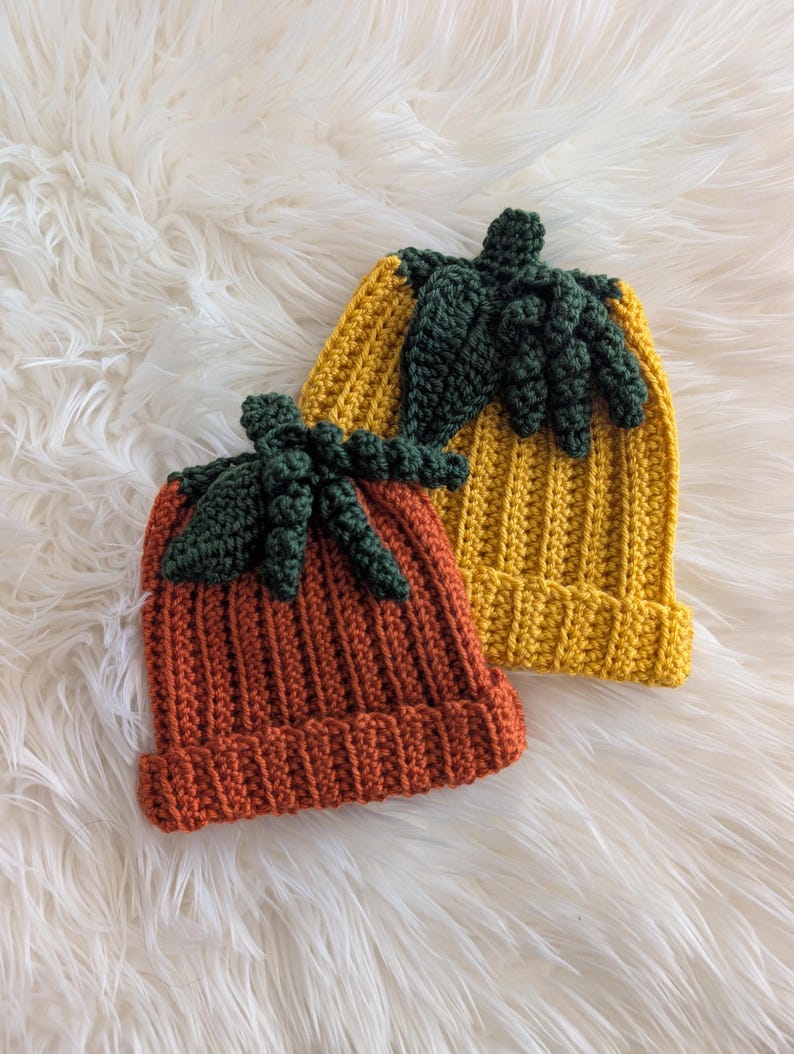 Puede incluir: Dos gorros de ganchillo hechos a mano con forma de calabaza. Uno es naranja, el otro amarillo. Ambos tienen detalles de hojas y tallos verdes. Los gorros son acanalados y se asientan sobre una superficie blanca y esponjosa.