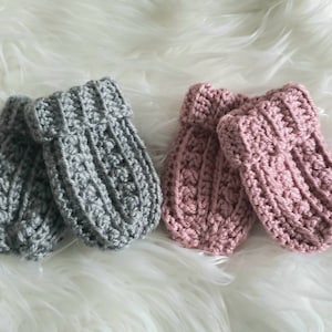 May include: Two pairs of handmade baby mittens. One pair is gray, the other is pink. The mittens are made of a textured, knitted material. The cuffs are ribbed. The mittens are displayed on a fluffy, white surface.