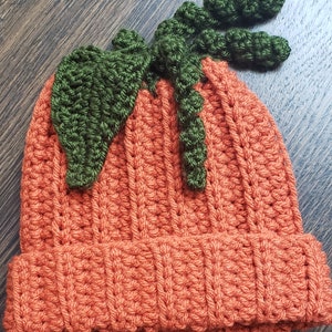 Puede incluir: Un gorro naranja tejido a ganchillo con un dise&ntilde;o de hoja y tallo verdes. El gorro est&aacute; hecho de un hilo texturizado y tiene un borde acanalado. La hoja y el tallo verdes est&aacute;n unidos a la parte superior del gorro.