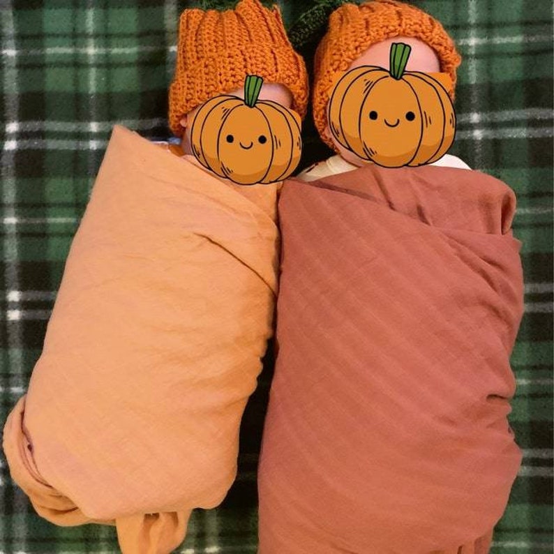 Puede incluir: Dos beb&eacute;s envueltos en mantas de color naranja y &oacute;xido, cada uno con un gorro de calabaza tejido de color naranja. Las caras de los beb&eacute;s est&aacute;n cubiertas con un gr&aacute;fico de calabaza de dibujos animados, creando un tema oto&ntilde;al festivo.