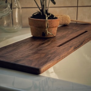 Puede incluir: Una bandeja de baño de madera marrón oscuro con una ranura larga y estrecha en el centro. La bandeja está sobre una bañera de porcelana blanca.