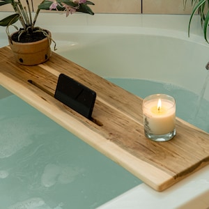 May include: A wooden bath tray with a phone slot, a lit candle, and a potted plant. The tray is floating in a bathtub filled with water.