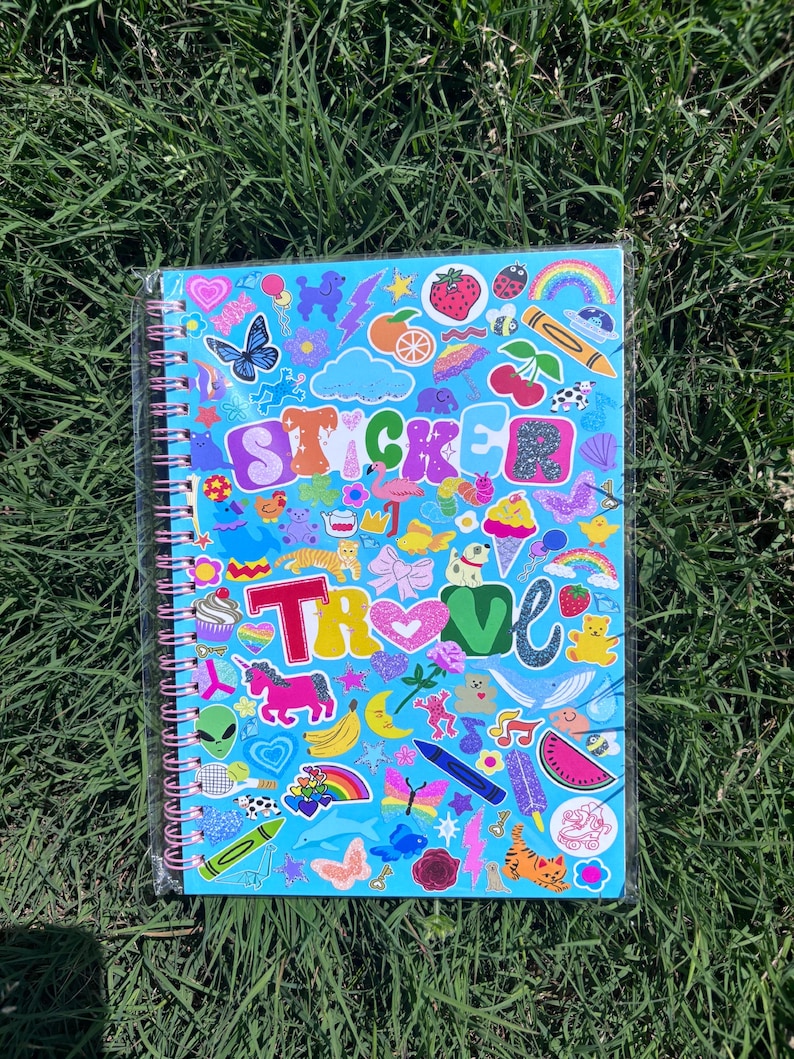 Puede incluir: Un cuaderno de espiral con una cubierta azul llena de pegatinas coloridas. Las pegatinas incluyen una mariposa, un arco&iacute;ris, una fresa y la palabra "STICKER". El cuaderno est&aacute; sobre hierba verde.