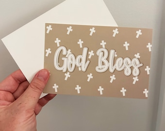 May include: A light brown card with a white background pattern of crosses. The card has the text "God Bless" in white with a white outline.