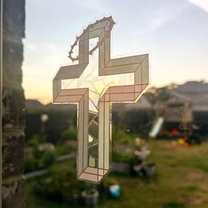 May include: A sticker of a geometric cross with a crown of thorns on a glass window. The cross is made of white and gold geometric shapes and is set against a blurred background of a yard.