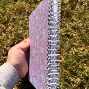 May include: A purple spiral bound notebook with a white floral pattern on the cover. The notebook has a silver wire binding.