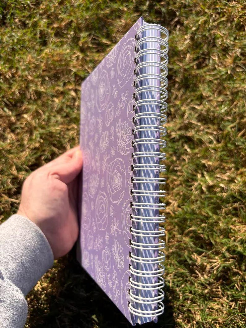 May include: A purple spiral bound notebook with a white floral pattern on the cover. The notebook has a silver wire binding.