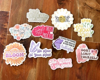Puede incluir: Una colección de 10 pegatinas coloridas con frases inspiradoras y diseños florales. Las pegatinas presentan frases como "Confía en el proceso", "Vales todo", "No te limites a ti mismo", y "El mundo es tuyo".