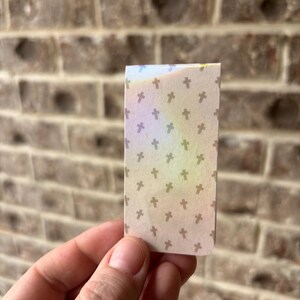 May include: A small, rectangular piece of paper with a pattern of small, brown crosses on a beige background. The paper has a rainbow iridescent sheen.