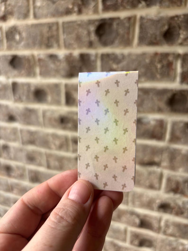 May include: A small, rectangular piece of paper with a pattern of small, brown crosses on a beige background. The paper has a rainbow iridescent sheen.