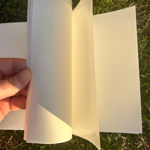 Puede incluir: Un cuaderno de bocetos abierto, con p&aacute;ginas color crema. Las p&aacute;ginas se est&aacute;n pasando, revelando la superficie en blanco. El libro se muestra sobre un fondo de hierba verde, lo que sugiere un entorno al aire libre. El papel parece grueso y de alta calidad.
