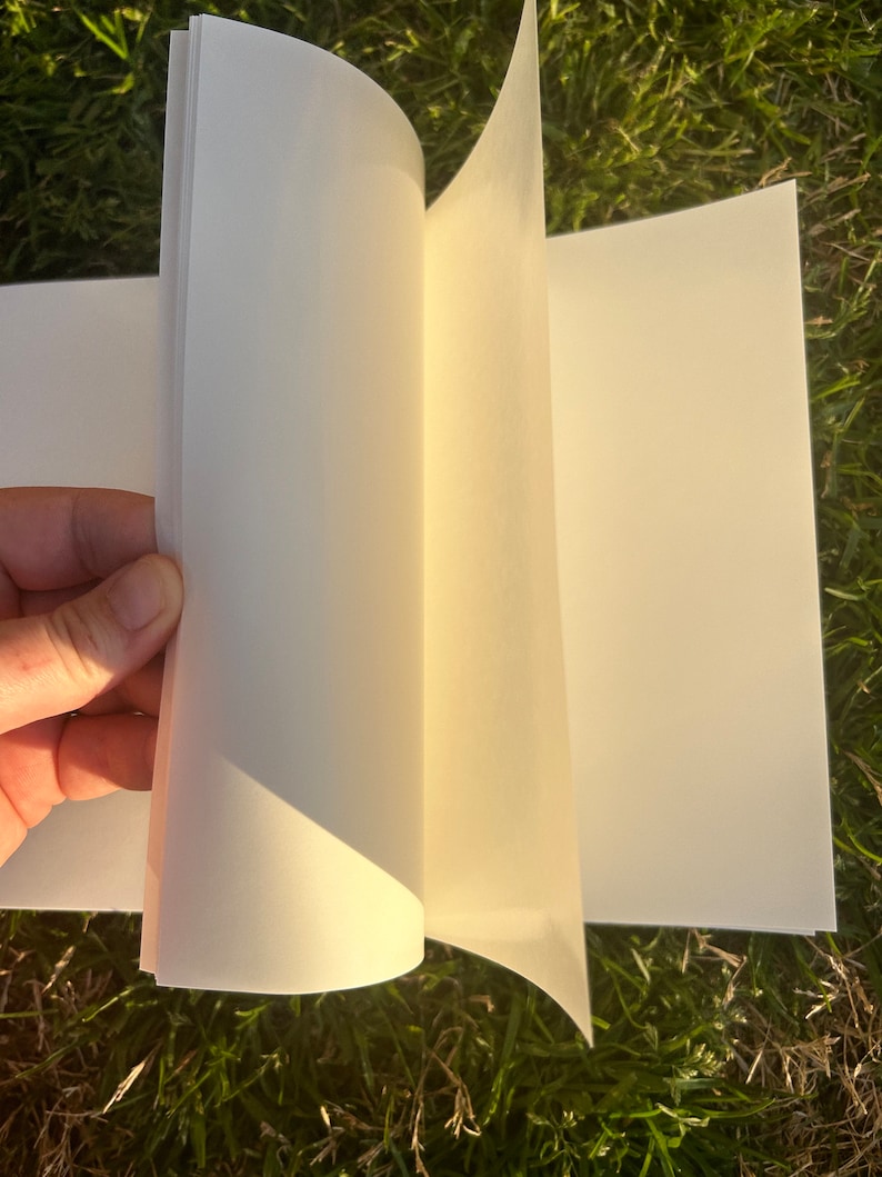 Puede incluir: Un cuaderno de bocetos abierto, con p&aacute;ginas color crema. Las p&aacute;ginas se est&aacute;n pasando, revelando la superficie en blanco. El libro se muestra sobre un fondo de hierba verde, lo que sugiere un entorno al aire libre. El papel parece grueso y de alta calidad.