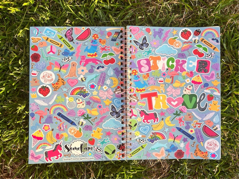 Puede incluir: Cuaderno espiral abierto con una cubierta azul claro llena de pegatinas coloridas. Las pegatinas incluyen mariposas, arco&iacute;ris, frutas, animales y las palabras "STICKER" y "TRUE LOVE". El cuaderno est&aacute; abierto sobre hierba verde.