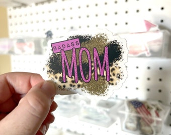 May include: A sticker with a gold glitter background and black and leopard print accents. The sticker says "BADASS MOM" in pink letters.