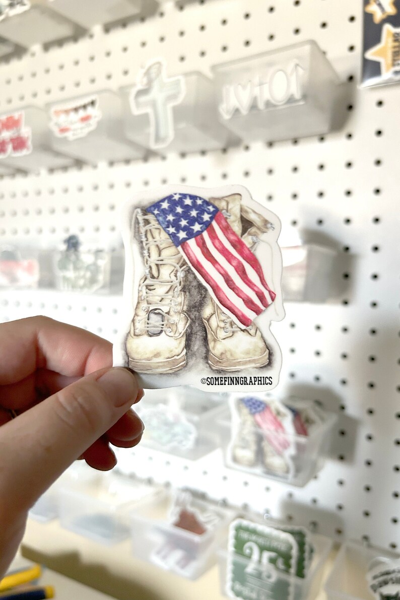 May include: A sticker featuring a pair of tan military boots with an American flag draped over them. The sticker has the text "@SOMEFINNGRAPHICS" at the bottom.