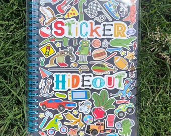 May include: A spiral-bound notebook with a black cover, filled with colorful stickers. The stickers feature various designs, including a guitar, dinosaur, and the words "STICKER HIDEOUT". The notebook is surrounded by green grass.