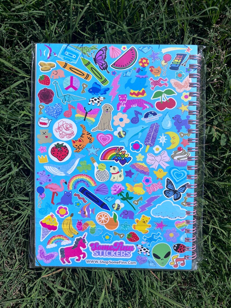 Puede incluir: Un cuaderno de espiral con una cubierta azul claro, densamente cubierto de pegatinas coloridas. Las pegatinas incluyen un arco&iacute;ris, un unicornio, mariposas y varios otros dise&ntilde;os lindos. El texto "Some Finn Stickers" y una direcci&oacute;n web son visibles.