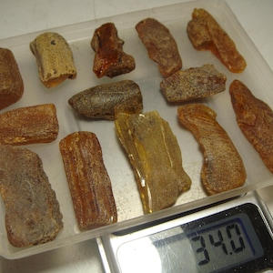 Puede incluir: Una colección de trece piezas de ámbar en bruto en varios tonos de marrón, naranja y amarillo, dispuestas en una bandeja de plástico transparente sobre una báscula digital que muestra un peso de 34,0 gramos. Las piedras tienen formas irregulares.