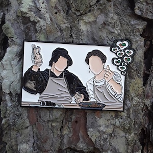 Puede incluir: Un pin de esmalte blanco con dos personas que llevan delantales y sostienen utensilios de cocina. El pin tiene un contorno negro y una nube en forma de corazón sobre las personas con corazones verdes y marrones.