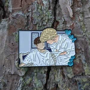 Könnte beinhalten: Eine weiße Emaille-Anstecknadel mit einer Cartoon-Illustration von zwei Personen in einem weißen Raum. Die Person links trägt ein weißes Hemd und die Person rechts trägt ein weißes Hemd und einen weißen Hut. Die Anstecknadel hat einen blauen Umriss und blaue Herzen um das Bild herum.