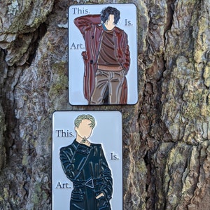 Op de afbeelding: Twee emaillepins met een gestileerde illustratie van een persoon in een jas. De pins zijn wit met zwarte tekst die "This is Art." luidt.