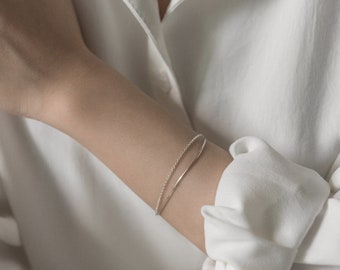 May include: Two silver bracelets on a person's wrist. The bracelet on top is a delicate chain, and the bracelet on the bottom is a simple band.