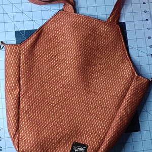 May include: A reddish-brown tote bag with a gold diamond pattern. The bag has two handles and a small black label with white text. The bag is made of a textured fabric.