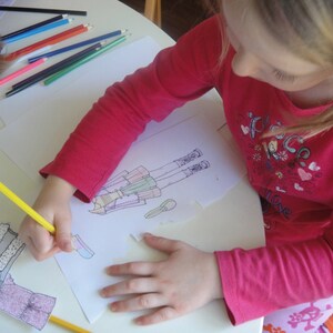 May include: A young person is coloring a drawing of a person wearing a dress and boots. The drawing is on a white sheet of paper. The person is using a yellow pencil to color the drawing. There are many colored pencils on the table.