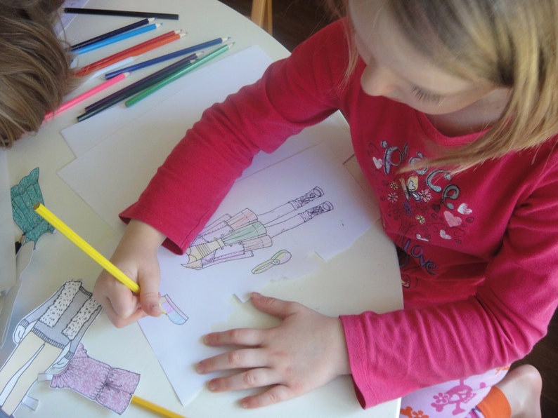 May include: A young person is coloring a drawing of a person wearing a dress and boots. The drawing is on a white sheet of paper. The person is using a yellow pencil to color the drawing. There are many colored pencils on the table.