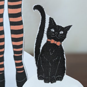 May include: A black cat with a red bow tie sits on a white surface. The cat has a long tail and is looking to the right.