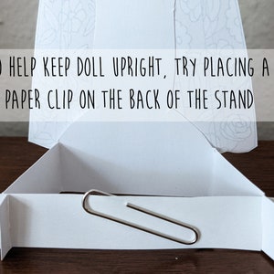 May include: A white cardboard doll stand with a paper clip attached to the back to help keep a doll upright. The text "TO HELP KEEP DOLL UPRIGHT, TRY PLACING A PAPER CLIP ON THE BACK OF THE STAND" is printed on the front of the stand.