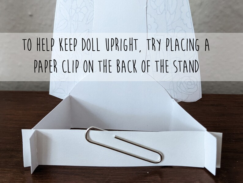 May include: A white cardboard doll stand with a paper clip attached to the back to help keep a doll upright. The text "TO HELP KEEP DOLL UPRIGHT, TRY PLACING A PAPER CLIP ON THE BACK OF THE STAND" is printed on the front of the stand.