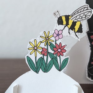 May include: A paper craft featuring a bee flying over a cluster of yellow and red flowers with green leaves. The bee is black and yellow with white wings. The flowers are set against a white background.