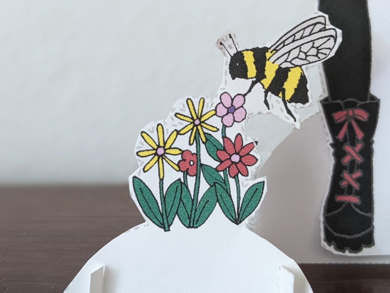May include: A paper craft featuring a bee flying over a cluster of yellow and red flowers with green leaves. The bee is black and yellow with white wings. The flowers are set against a white background.