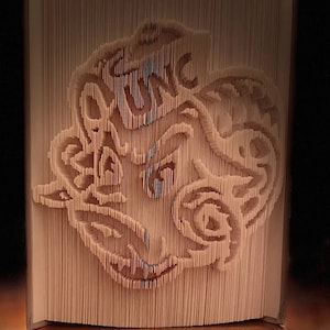 May include: A book art sculpture of a stylized animal face, created by folding the pages of a book. The sculpture is made of cream-colored paper and has a detailed design.