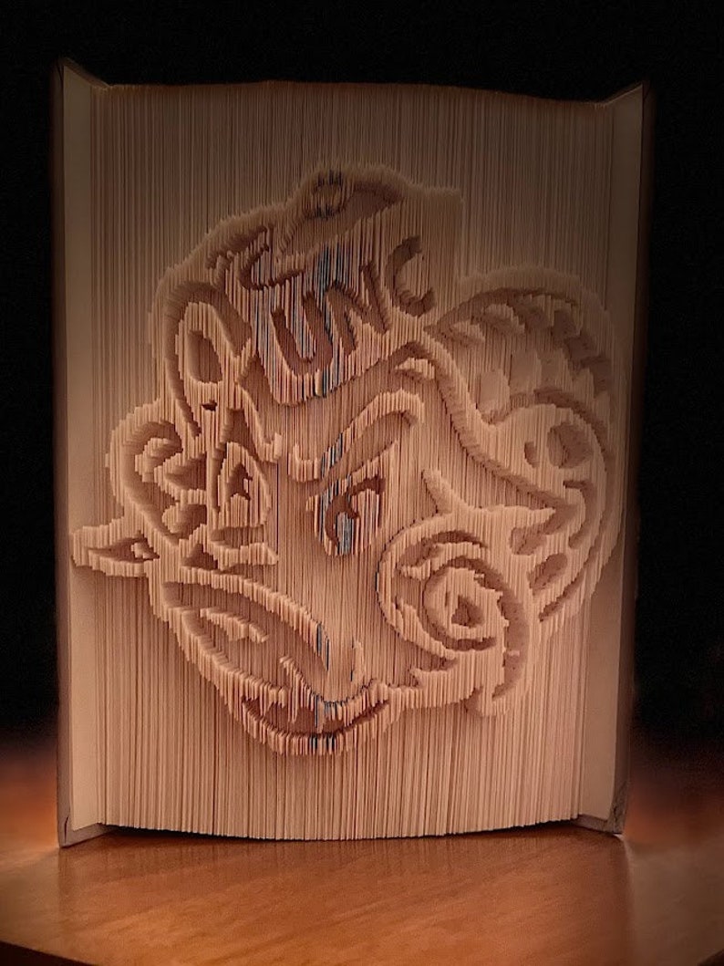 May include: A book art sculpture of a stylized animal face, created by folding the pages of a book. The sculpture is made of cream-colored paper and has a detailed design.