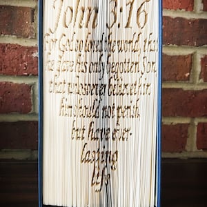 Puede incluir: Un libro azul con un diseño en forma de corazón blanco en la portada. El corazón está hecho de páginas dobladas y el texto "Juan 3:16" es visible en la portada. El texto "Porque tanto amó Dios al mundo, que dio a su Hijo unigénito, para que todo aquel que en él cree, no se pierda, mas tenga vida eterna" es visible en las páginas dobladas.