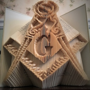 Könnte beinhalten: Ein Buch, das zu einer dreidimensionalen Skulptur einer Rose und eines Zirkels und eines Geodreiecks gefaltet ist, ein Symbol der Freimaurerei.