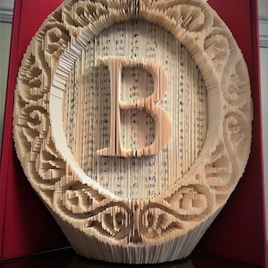 May include: A book art sculpture of the letter "B" in a circular frame. The sculpture is made from the pages of a book that have been folded and cut to create a three-dimensional design. The sculpture is displayed on a wooden surface.