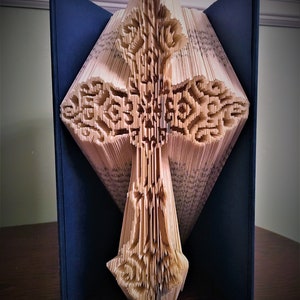 May include: A book with pages folded into a three-dimensional cross shape. The cross is made of paper and has a detailed design. The book is open and the cross is standing upright. The book is blue and the cross is beige.