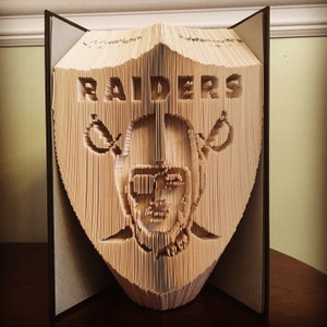 May include: A unique book art sculpture featuring the Las Vegas Raiders logo and the word "RAIDERS" cut into the pages. The sculpture is displayed open, showcasing the intricate details of the team's emblem.