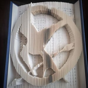 May include: A book with pages meticulously folded to create a three-dimensional design. The design features a circular shape with a stylized bird or emblem in the center. The book's edges are blue, and the pages are a light beige color.