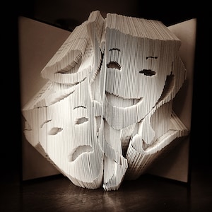 May include: A book folded into the shape of two faces, one smiling and one frowning. The faces are made of white paper and are set against a dark background.