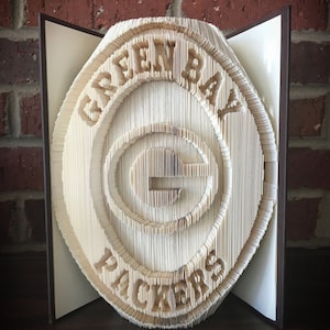 May include: A handcrafted book sculpture featuring the Green Bay Packers logo. The logo, including the words "GREEN BAY PACKERS" and the "G" symbol, is carved into the pages of an open book, creating a 3D effect. The book is displayed against a brick wall.