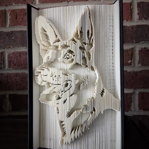 Può includere: Una scultura di arte libraria di una testa di cane, creata piegando le pagine di un libro. La scultura è realizzata in carta bianca ed è esposta su uno sfondo di mattoni.