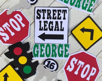Puede incluir: Un conjunto de decoraciones para una fiesta de cumpleaños número 16 con una señal de alto, una señal de "Street Legal" con una flecha, un semáforo, una llave y un letrero que dice "George".